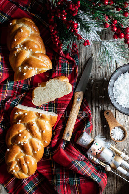 Top view of wooden table with cut traditional braided bread and knife placed on checkered Christmas tablecloth with decorative objects — Stock Photo