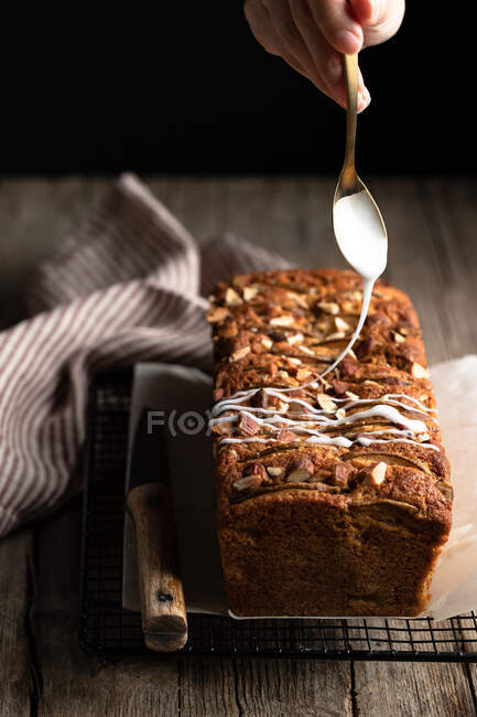 Crop housewife with spoon in hand pouring white sugar icing over homemade banana bread with nuts placed on metal grid on wooden table — Stock Photo