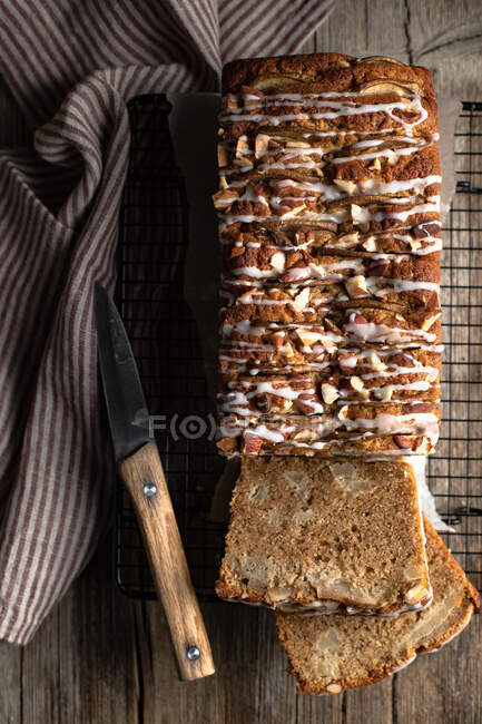 Top view of sliced appetizing homemade banana bread with nuts and sugar icing placed on wooden table with knife and tablecloth — Stock Photo