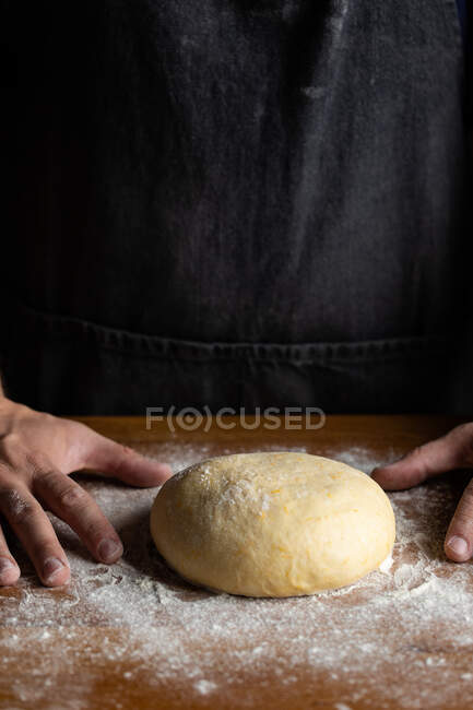 Кукурудзяний чоловічий пекар у чорному фартусі, що формує хлібний хліб, стоячи за дерев'яним столом, забитим білим борошном — стокове фото