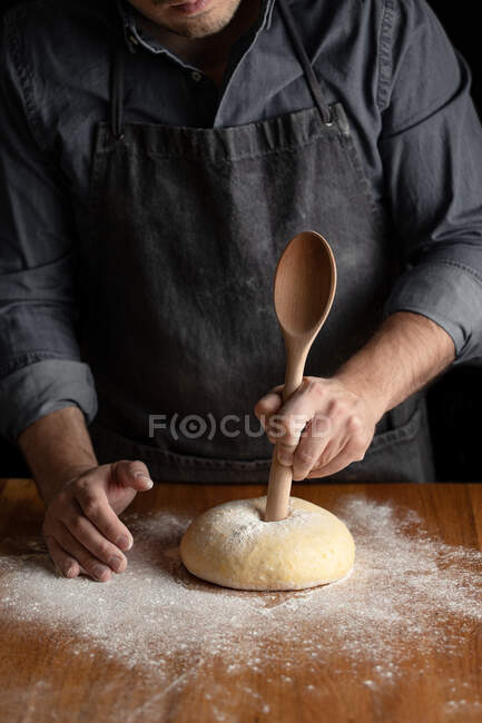 Crop male baker in black apron using big wooden spoon for making hole in dough while forming artisan round bread loaf at wooden table — Stock Photo