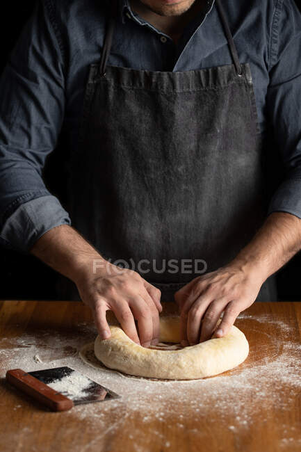 Мужчина-пекарь в черном фартуке делает дыру в тесте, формируя ручной круглый хлеб за деревянным столом — стоковое фото