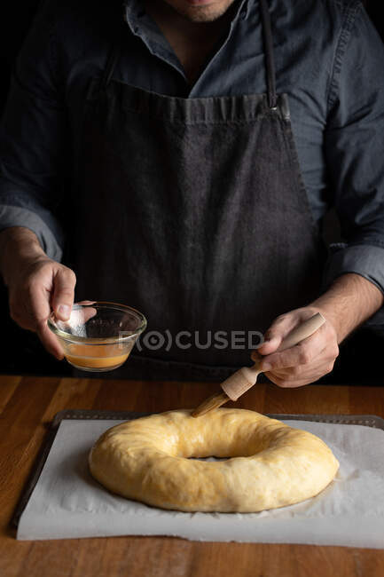 Ernte männlicher Koch in schwarzer Schürze schmiert ungebackenes rundes Brot mit Eigelb, während er am Holztisch steht — Stockfoto