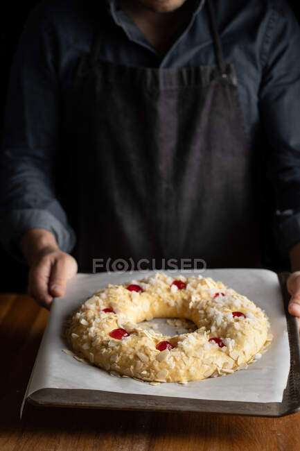 Шеф-повар в черном фартуке держит неиспеченный круглый хлеб, увенчанный вишней, стоя за деревянным столом. — стоковое фото