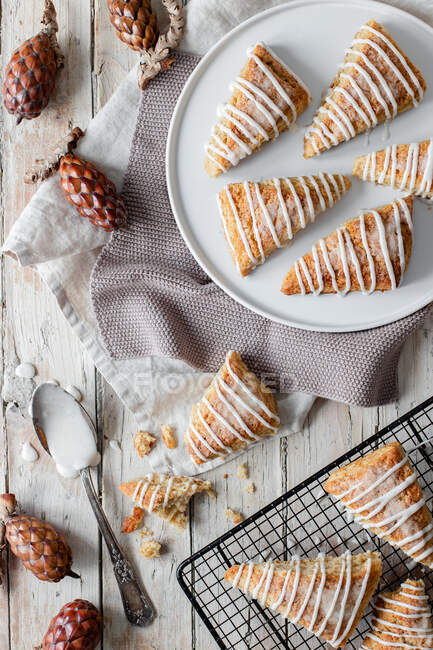 Top view of tasty sweet homemade triangle scones decorated with sugar icing placed on white plate and on metal grid on wooden table with decorative tree cones — Stock Photo