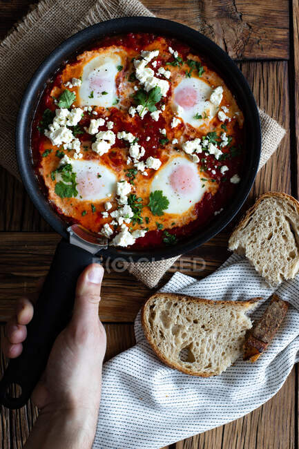 Top view of crop person putting on wooden table frying pan with delicious shakshuka made with fried eggs with vegetables and feta cheese — Stock Photo
