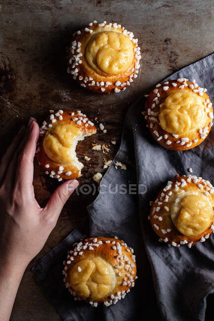Top view of crop female holding bitten bun while standing at table with freshly baked yummy sweet buns with cream and sprinkles — Stock Photo