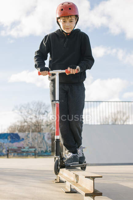 Adolescente activo en casco protector con patinete scooter de pie en rampa en skate park mientras se prepara para realizar truco en soleado día de primavera - foto de stock