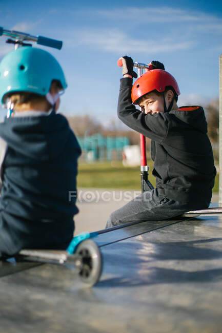 Side view of cheerful teen boy in helmet with kick scooter talking to unrecognizable friend while sitting together on ramp in skate park in sunny spring day — Stock Photo