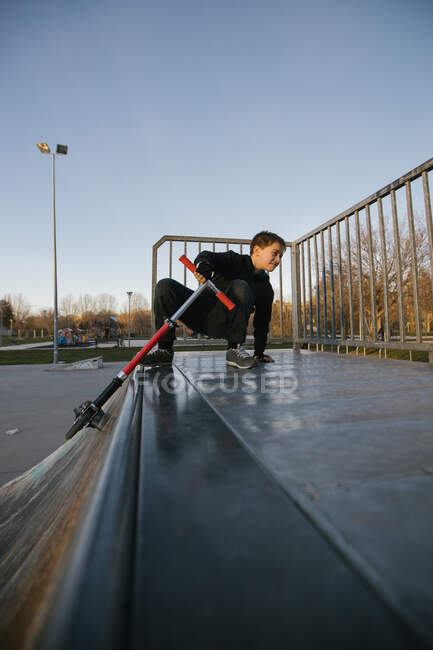 Активный подросток в защитном шлеме со скутером, сидящим на рампе в скейт-парке во время подготовки к выполнению трюка в солнечный весенний день — стоковое фото