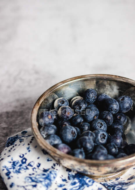 From above of fresh ripe blueberries in metal bowl placed on white and blue napkin on table against shabby wall — Stock Photo