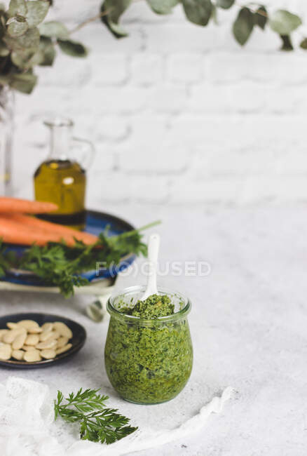 Frasco de vidro com delicioso molho pesto caseiro feito com folhas de cenoura colocadas na mesa com ingredientes contra a parede de tijolo branco com plantas verdes — Fotografia de Stock
