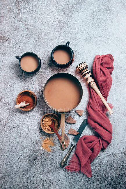 Vue de dessus du chocolat chaud épicé mexicain traditionnel avec de la cannelle et des poudres de poivron rouge servies en pot et des tasses placées avec des morceaux de chocolat et un fouet en bois avec un chiffon rouge sur fond gris — Photo de stock