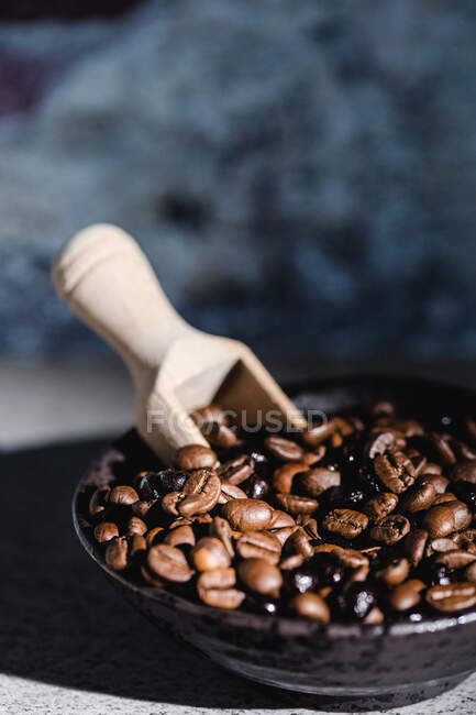 Desde arriba del tazón negro con granos de café tostados frescos aromáticos y cuchara de servir de madera colocada en la mesa con fondo borroso - foto de stock