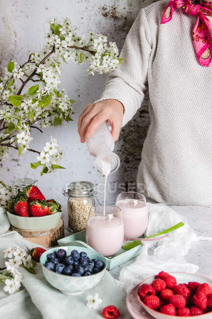 Mulher de colheita em roupas casuais derramando saboroso smoothie caseiro em vidro enquanto prepara o café da manhã saudável à mesa com frutas frescas e flores em casa — Fotografia de Stock