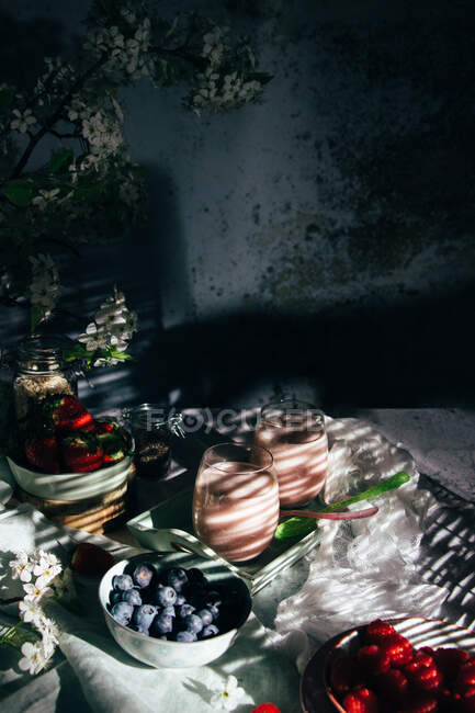 De acima mencionada composição de batido de morango são em copos colocados na mesa com várias bagas frescas e flores no quarto com luz solar e sombras — Fotografia de Stock