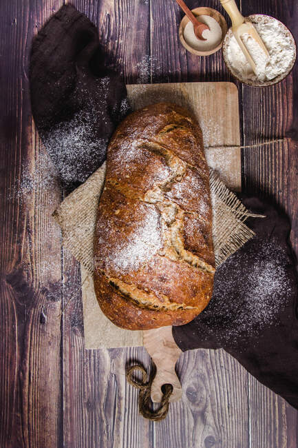 Vista superior de pão crocante apetitoso de pão recém-assado polvilhado com farinha colocada a bordo com guardanapos em mesa de madeira com ingredientes — Fotografia de Stock