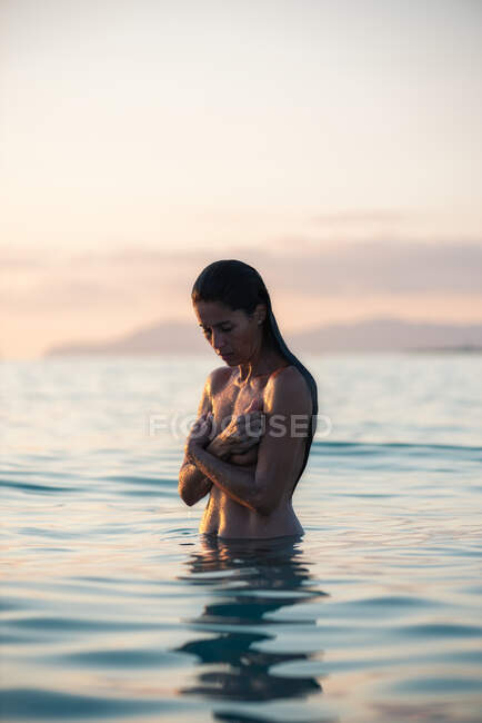 Mujer desnuda cubriendo el pecho mientras está de pie en el agua de mar ondulante contra el cielo al atardecer en la naturaleza - foto de stock