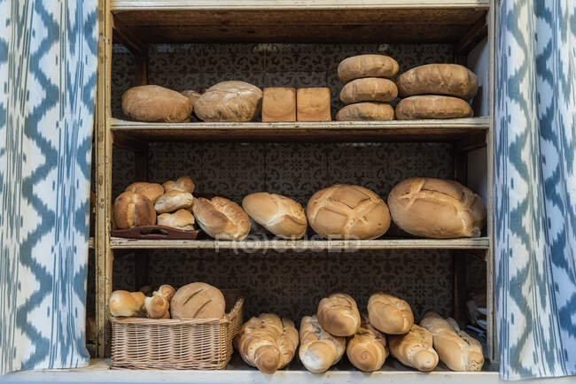 Laibe mit verschiedenen frischen Broten zum Verkauf in schäbigen Regalen in der Nähe von Gardinen in der Bäckerei platziert — Stockfoto