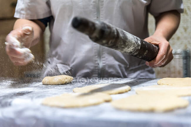 Panadero irreconocible con rodillo derramando harina en la mesa mientras prepara pastelería en la cocina - foto de stock