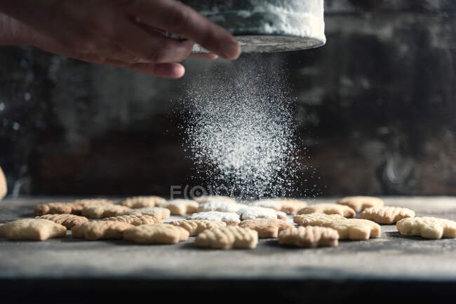 Panadero irreconocible usando tamiz para derramar azúcar en polvo en sabrosas galletas en la mesa en la cocina - foto de stock