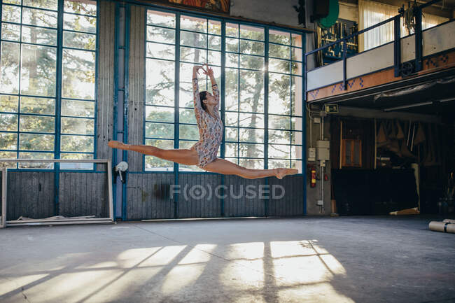Side view of young slim ballerina in gymnastic suit jumping while practicing dance movements in light spacious studio with large window — Stock Photo