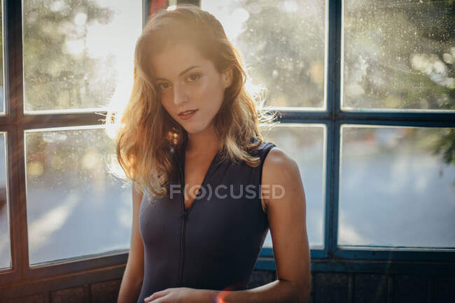 Attraktive junge Frau in schwarzem Kleid blickt in die Kamera, während sie im Gegenlicht gegen Fenster steht — Stockfoto