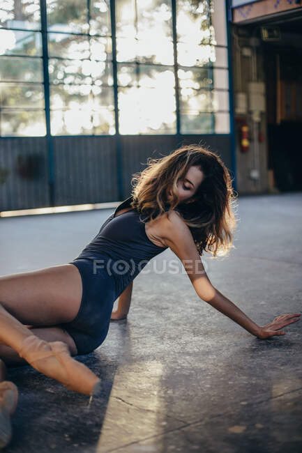 Вид сбоку на стройную молодую балерину в боди-костюме и пуантах, сидящую на полу и практикующую танцевальные движения во время тренировки в студии — стоковое фото