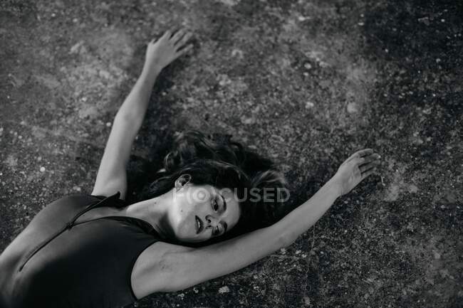 Черное и белое от соблазнительной расслабленной молодой женщины в телесном костюме, лежащей на потрепанной земле и смотрящей в сторону — стоковое фото