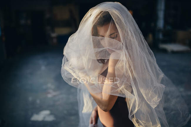 Young talented female dancer with closed eyes in bodysuit dancing with transparent veil while training alone in light studio — Stock Photo