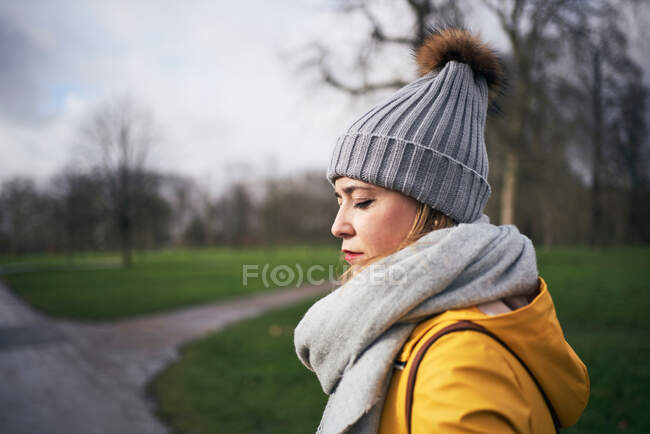 Side view of calm young female in trendy outerwear with scarf and hat standing in park with green grass and leafless trees — Stock Photo