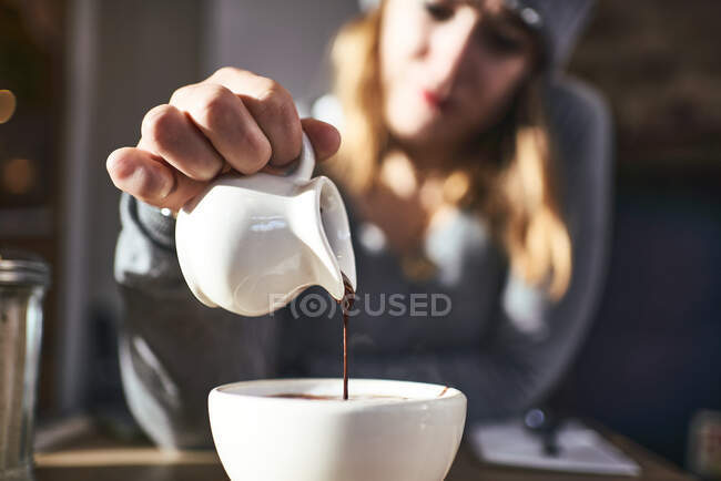 Размытая неузнаваемая женщина в теплом свитере и шляпе, наливающая шоколадный сироп в белую кружку с горячим напитком, сидя за столом в солнечном кафе — стоковое фото