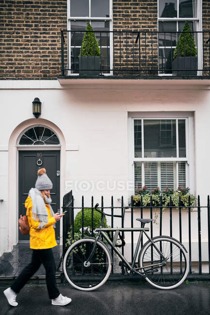 Seitenansicht einer Frau in warmer Oberbekleidung mit Rucksack und Smartphone beim Spazierengehen auf der Stadtstraße in der Nähe eines Altbaus mit traditioneller Architektur und im Herbst in der Nähe abgestelltem Fahrrad — Stockfoto