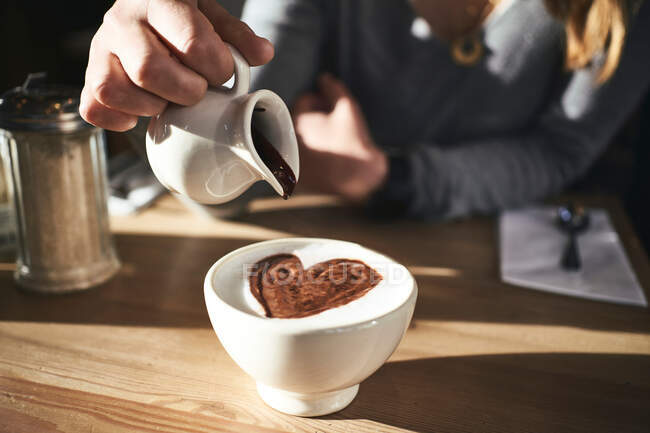 Cropped unrecognizable woman in warm sweater and hat pouring chocolate syrup into white mug with hot beverage while sitting at table in sunny cafe — Stock Photo