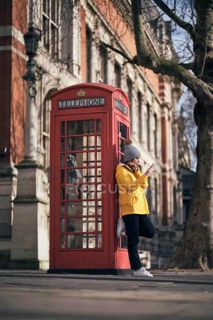 Vista lateral da mulher moderna em outerwear inclinado na caixa de telefone vermelha e smartphone de navegação enquanto está de pé na calçada na rua da cidade — Fotografia de Stock