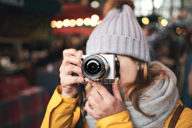 Unrecognizable female traveler in warm clothes taking pictures with camera while standing on city street with illumination in winter evening — Stock Photo