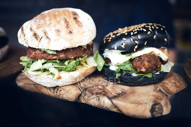 Deliciosos hambúrgueres brancos e pretos com alface verde e queijo servido em tábua de madeira em mesa preta — Fotografia de Stock