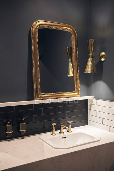 Дзеркало з золотою рамою, що висить на чорній стіні біля лампи над мармуровою лічильницею з раковиною в елегантній ванній кімнаті — стокове фото