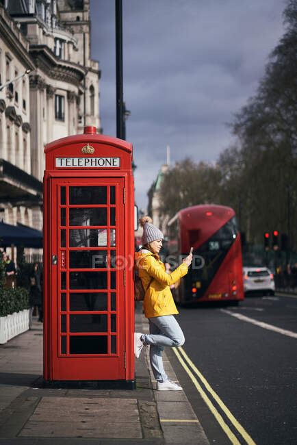 Vista lateral da mulher moderna em outerwear inclinado na caixa de telefone vermelha e smartphone de navegação enquanto está de pé na calçada na rua da cidade — Fotografia de Stock