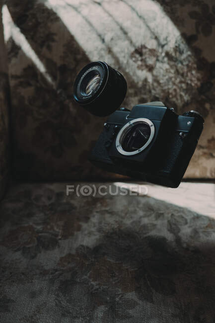 Câmera de foto quebrada e lente caindo no sofá macio com ornamento floral no quarto escuro — Fotografia de Stock