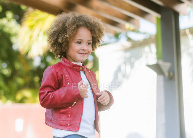 Чарівна маленька дівчинка з кучерявим волоссям у повсякденній сорочці посміхається, дивлячись далеко, стоячи на вулиці — стокове фото