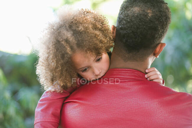 Вид ззаду на невпізнаваного етнічного чоловіка в червоній шкіряній куртці, що обіймає милу кучеряву волохату дочку, проводячи час разом у зеленому парку — стокове фото
