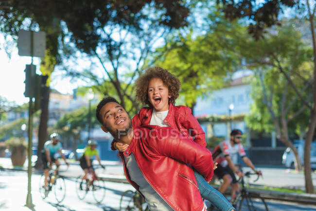 Happy young ethnic man giving piggyback ride to delighted little daughter while enjoying free time together on city street — Stock Photo