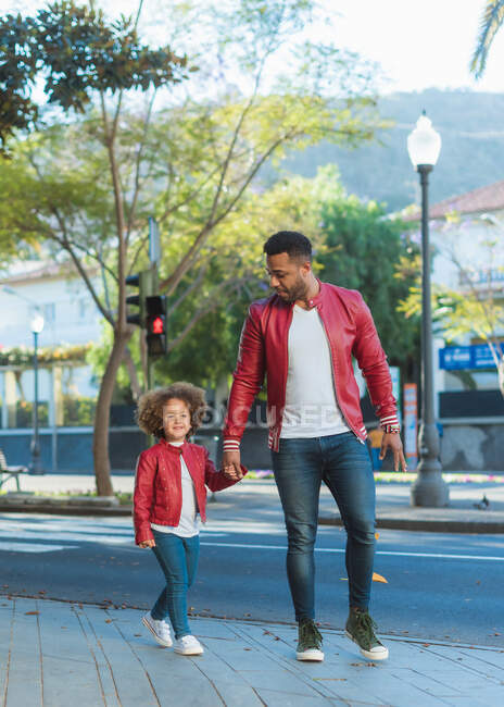 Щасливий молодий етнічний чоловік з маленькою дочкою, одягнений у схожий одяг, тримає руки під час прогулянки по міській вулиці в сонячний день — стокове фото