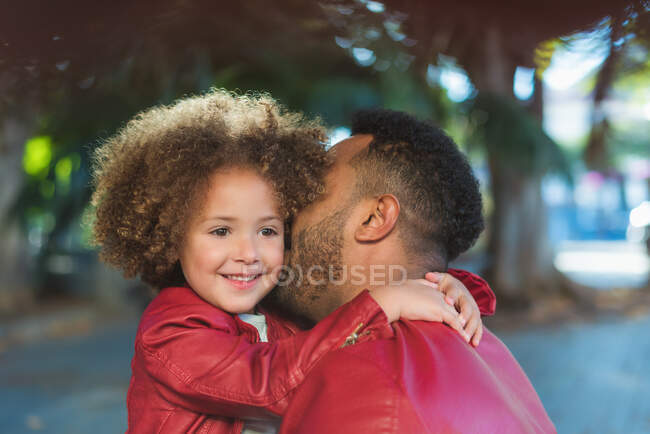 Vista lateral de adorable chica étnica alegre abrazando padre feliz con chaqueta de cuero similar mientras descansan juntos en el parque en el día soleado - foto de stock