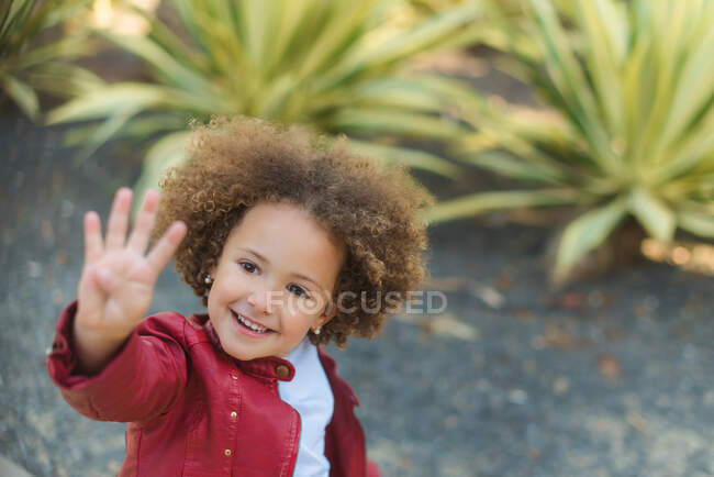 Високий кут весела маленька кучерява дівчинка в червоній куртці, що показує чотири пальці і посміхається, стоячи проти зелених тропічних рослин в парку — стокове фото