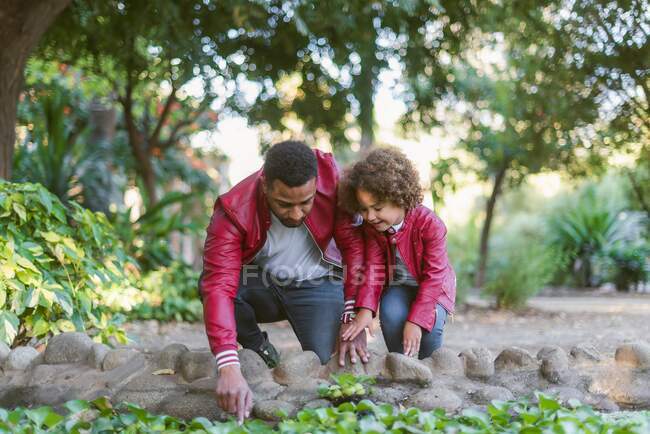 Молодой человек и маленькая девочка в одинаковых кожаных куртках и джинсах наблюдают за рыбой в пруду, отдыхая вместе в зеленом парке — стоковое фото