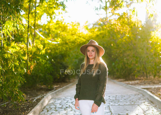 Positive young female in stylish outfit and hat looking at camera while standing on stone pathway in green park in sunny day — Stock Photo