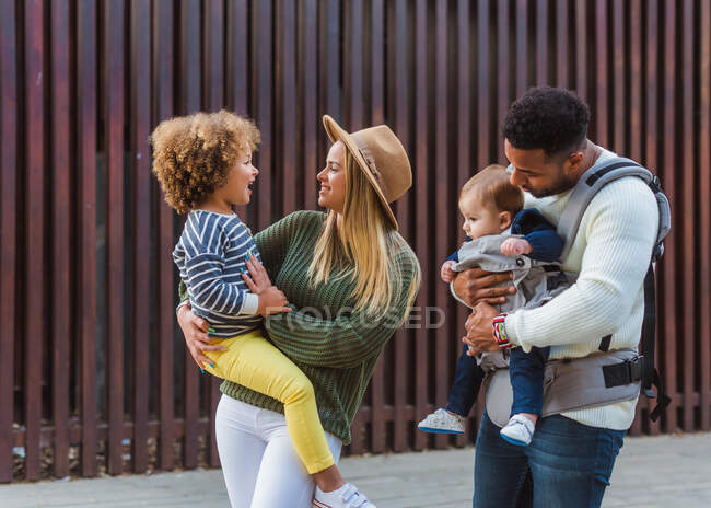 Alegre elegante joven pareja multirracial con hijo pequeño y pequeña hija en ropa casual caminando juntos a lo largo de valla de madera en la calle de la ciudad - foto de stock
