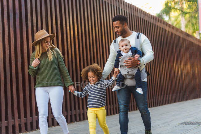 Cheerful stylish young multiracial couple with infant son and little daughter in casual wear walking together along wooden fence on city street — Stock Photo